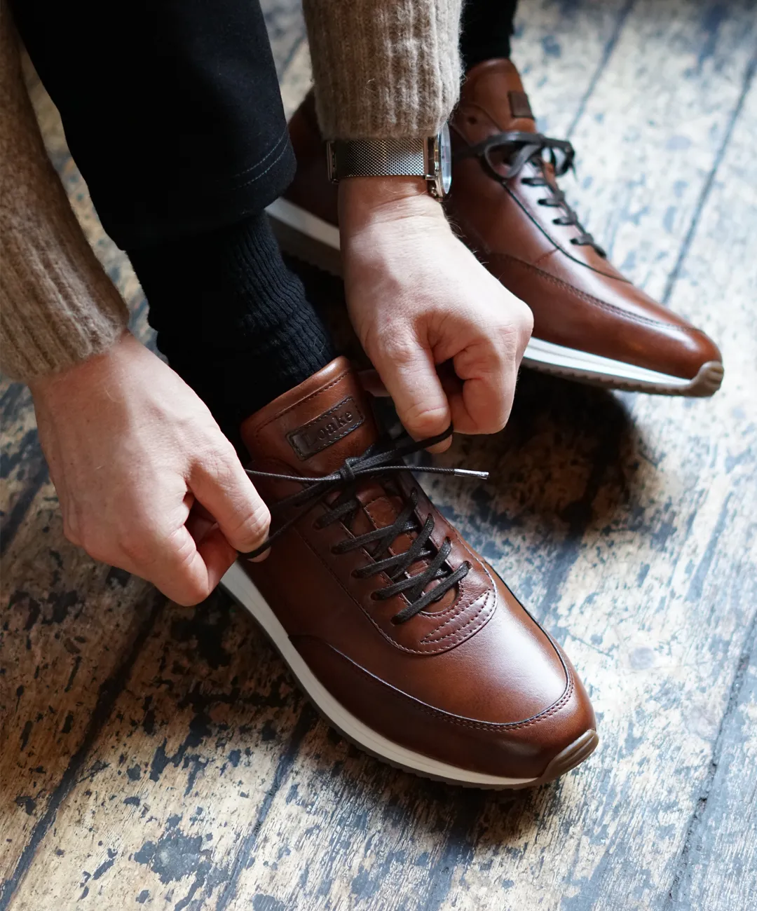 Man tying up his trainers in a public setting. Shoes shown are Bannister in cedar calf